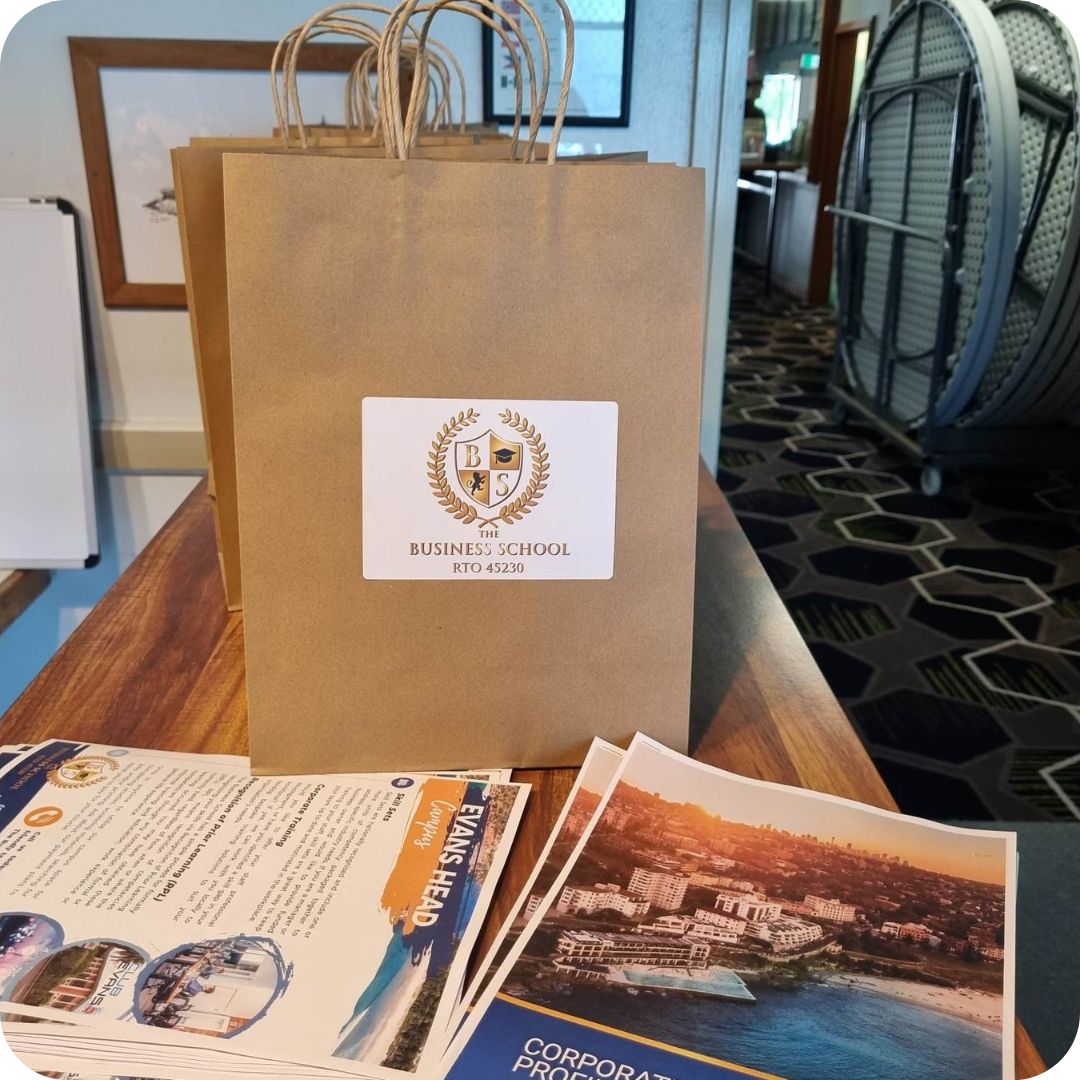 A brown paper bag with a white label of a business school on a table with brochures at the Evans Head Campus